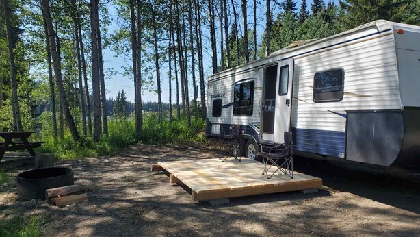 A travel trailer with a wooden deck, a picnic table, and a campfire setup in a forested area. - Trailer on Spacious lakefront Campground (Lone Butte)