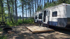Una caravana de viaje con una terraza de madera, una mesa de picnic y una zona para fogata en un área boscosa.