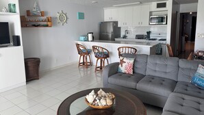 Una sala de estar moderna con un sofá gris, una mesa de centro con conchas marinas, sillas de mimbre, una zona de cocina con armarios blancos y un ventilador de techo.