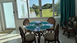 Un comedor con una mesa redonda, cuatro sillas, un ventilador de techo y vistas a un jardín a través de grandes ventanales.