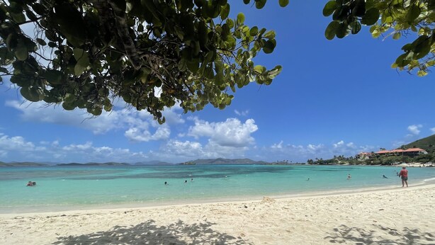 Una playa con aguas turquesas cristalinas, arena blanca y colinas al fondo.
