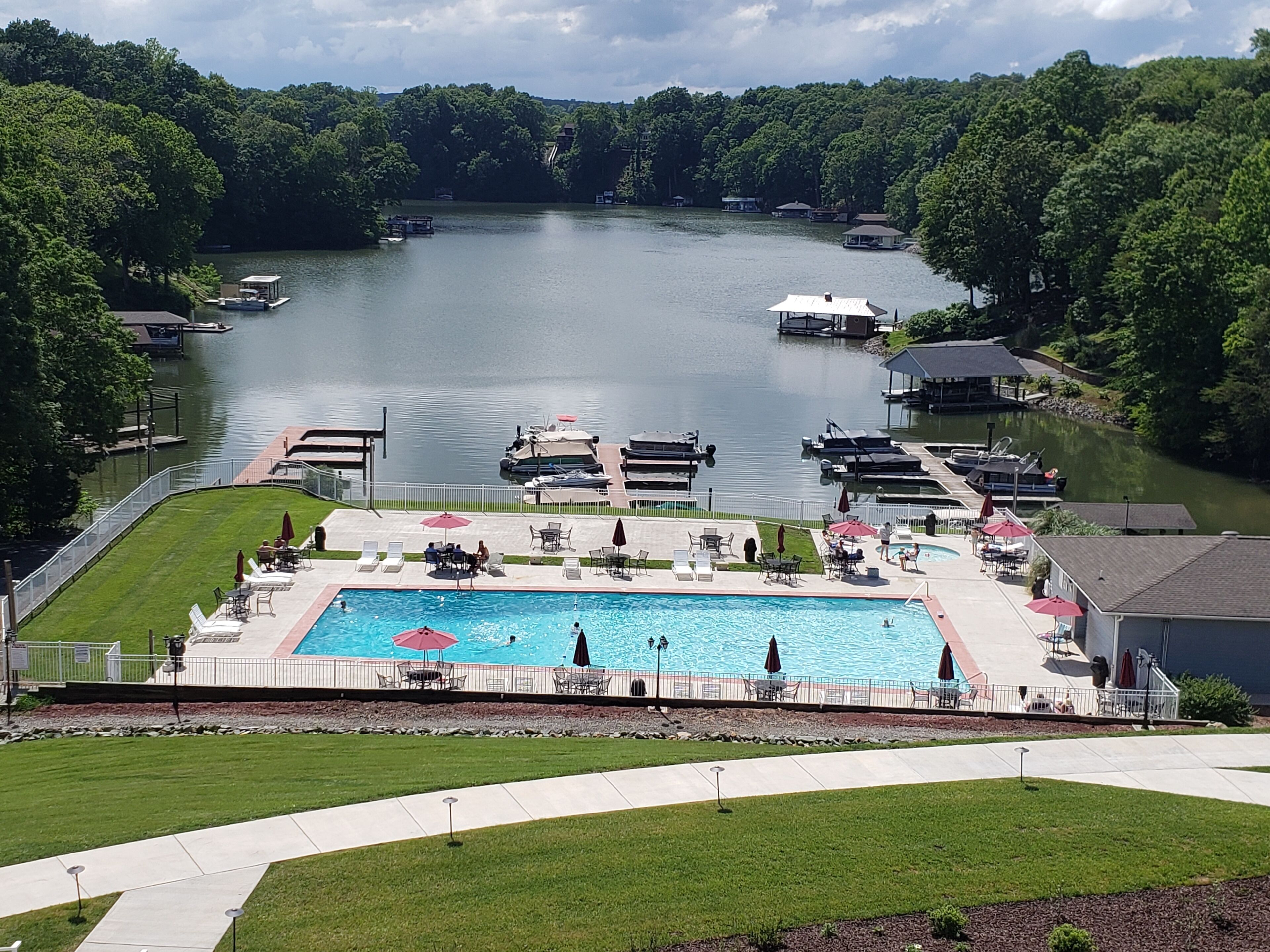 Una piscina con sillones de descanso y sombrillas, rodeada por una terraza y vistas a un lago con barcos y casas amarradas.