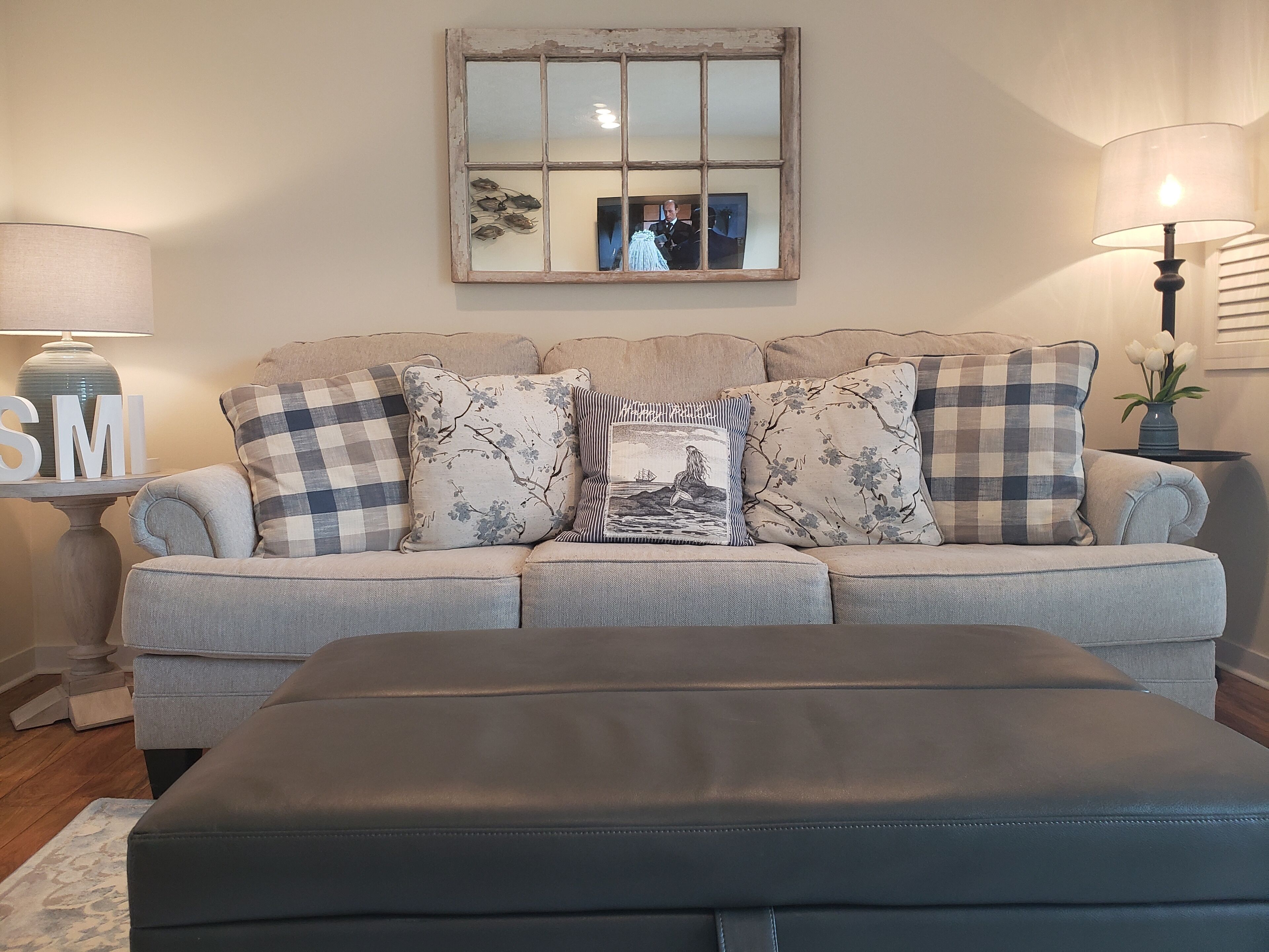 A living room with a grey sofa, multiple patterned cushions, a black ottoman, a wooden side table with a lamp, and a mirror on the wall.