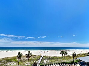 A beach view with a wooden boardwalk, palm trees, and beach umbrellas. - Windward Pointe 403 – Gulf-Front Retreat in Orange Beach! (Orange Beach)