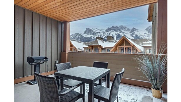 A balcony with a view of snow-covered mountains, a table with chairs, and a grill. - Rundle Rock Retreat | Majestic Views - Luxury Condo (Canmore)