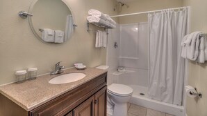 A bathroom with a round mirror, two sinks, a toilet, a shower with a curtain, and neatly folded towels on a rack. - Oceanfront One Bedroom Suite, Stunning Views & King Bed! (Lincoln City)