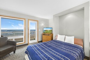 A hotel room with a large bed, a TV, a chair, a dresser, and a balcony with a view of the ocean.