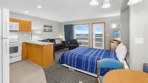 Habitación de hotel pequeña con kitchenette, una cama con colcha azul estampada, una silla, un escritorio y un televisor.