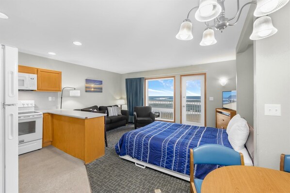 A compact hotel room with a kitchenette, a bed with a blue patterned bedspread, a chair, a desk, and a television.
