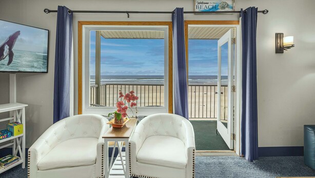 Una habitación con dos sillones blancos, una mesita con un florero de flores rosas, un televisor en la pared y un balcón con vista al océano.