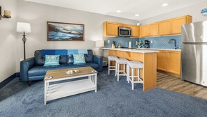 Un espacio habitable compacto con kitchenette, un sofá azul con cojines, una mesa de centro y un cuadro en la pared.