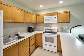 A compact kitchen with wooden cabinets, a white countertop, a built-in microwave, a stove, and a coffee maker. - Beachfront Suite - Ocean Views - Heated Pool & Hot Tub! (Lincoln City)
