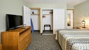 A hotel room with a television on a wooden dresser, a wardrobe with hanging clothes, a chair, and a bed with patterned bedding.