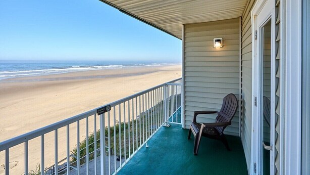 A balcony with a view of the beach, a chair, and a small table.
