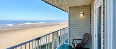 A balcony with a view of the beach, a chair, and a small table.
