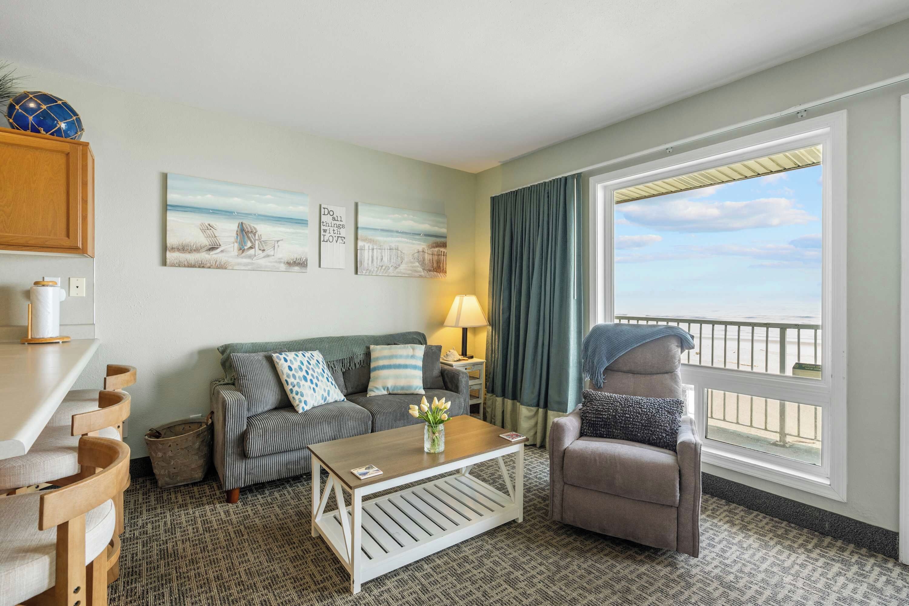 A hotel room with a balcony, a sofa, a chair, a coffee table, and artwork on the wall.