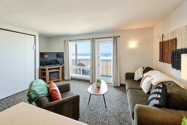 A hotel room with a balcony, a TV, a sofa, a small table, and a view of the ocean.