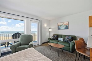 Een kamer met een groene bank, een houten bijzettafeltje, een bed met een beddeken met patroon, een stoel en uitzicht op het strand door een groot raam.