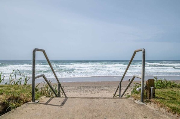 A concrete pathway leading to a beach with metal railings and a wooden post on the right.