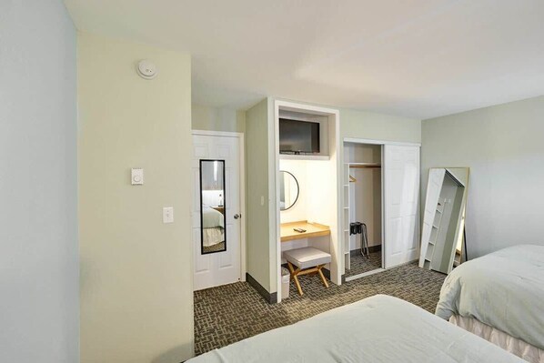 A hotel room with a bed, a desk with a chair, a mirror, a wardrobe, and a door leading to another room.