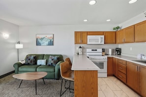 A compact kitchen with wooden cabinets, a microwave, an oven, and a small dining area with a green sofa and chairs. - Oceanfront Suite - Sleeps Six - Full Kitchen - Pool and Hot Tub! (Lincoln City)