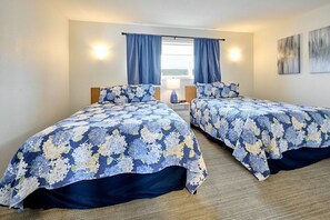Ein Doppelzimmer mit zwei Betten, jeweils mit floralem Bettbezug, einem Nachttisch mit Lampe und einem Fenster mit Vorhängen.