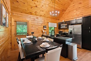 A wooden cabin interior with a dining table set for four, a kitchen with modern appliances, and a chandelier.