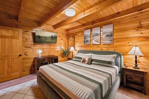A wooden cabin bedroom with a bed, two bedside tables, a lamp, a mirror, and three paintings on the wall.