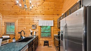 A compact kitchen with wooden walls, a stainless steel refrigerator, a sink, and a window with a view of greenery. - King Beds, Secluded Deck, Hot Tub, Outdoor Pool, Game Room, Pool Table (Sevierville)
