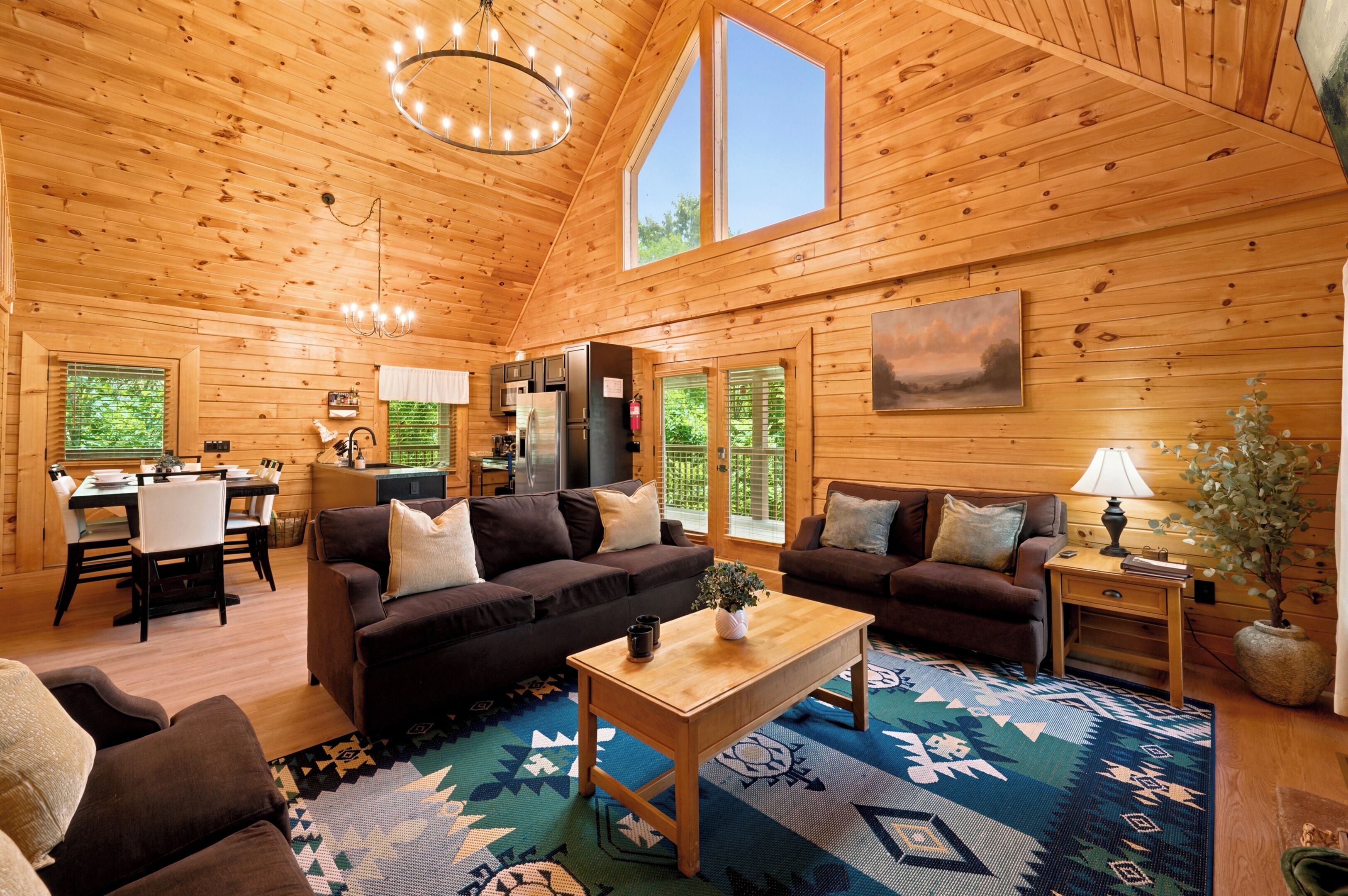 A wooden cabin interior with a dining area, two sofas, a coffee table, and a large window.