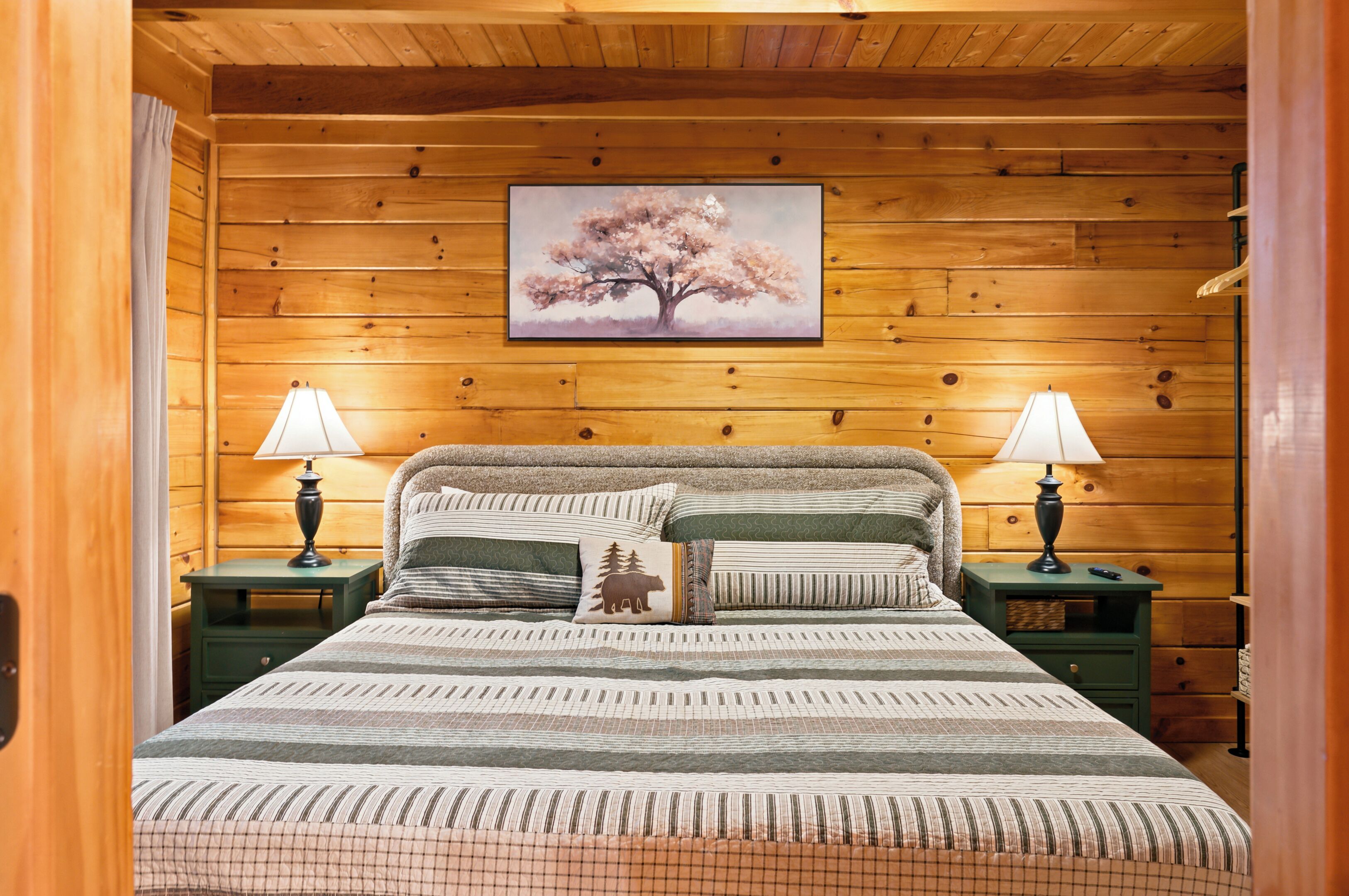 A wooden cabin bedroom with a bed, two nightstands, and a painting of a tree.