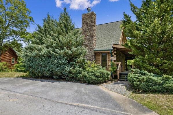 A log cabin with a stone chimney, surrounded by dense evergreen trees and a gravel driveway. - King Beds, Secluded Deck, Hot Tub, Outdoor Pool, Game Room, Pool Table (Sevierville)