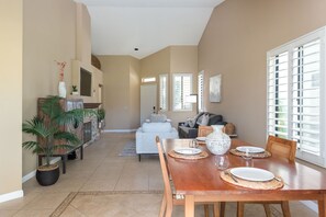 Amplia sala de estar con mesa de comedor preparada para cuatro, chimenea y ventana grande con persianas.