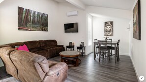 Un salon moderne avec un canapé en cuir marron, une table basse ronde, un téléviseur à écran plat, une table à manger avec des chaises et un grand tableau représentant une forêt, accroché au mur.