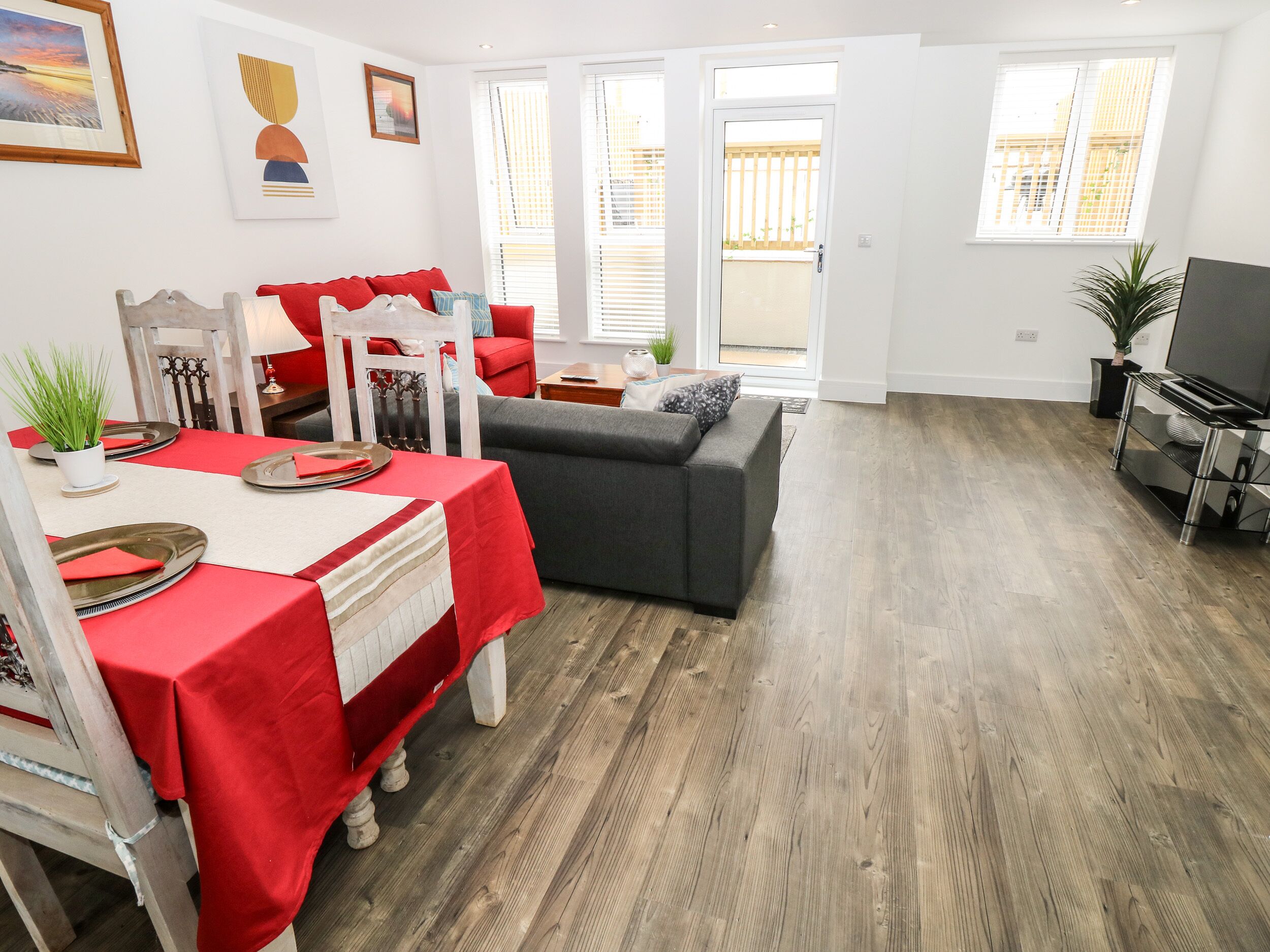 A modern living room with a grey sofa, a dining table with red tablecloths, and a TV stand.