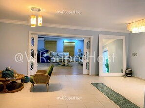 Una sala de estar moderna con sofá, sillas, una mesa de centro y una mesa de comedor. Hay un separador de vidrio que da a otra habitación.