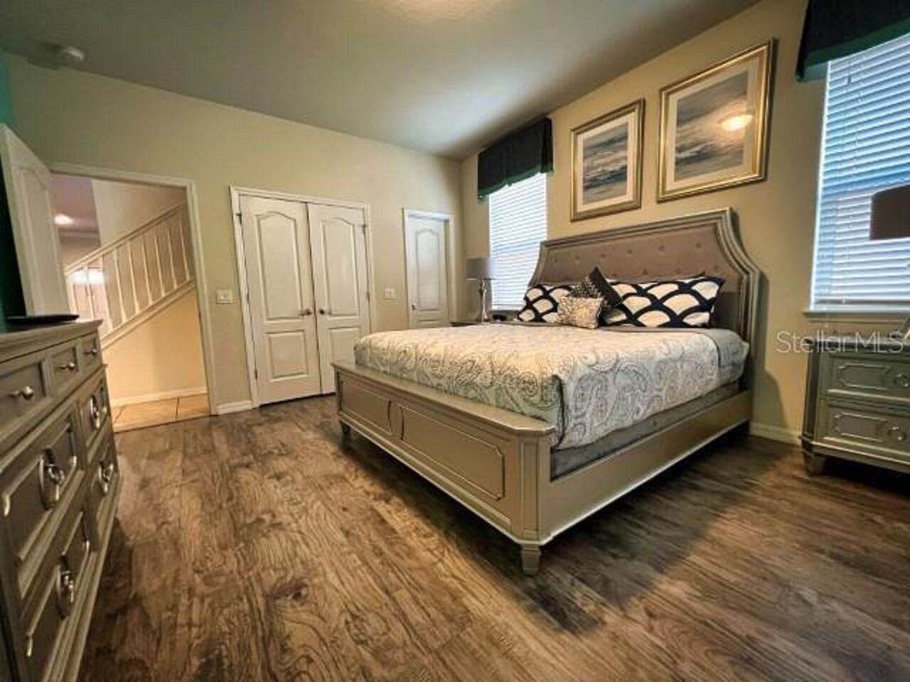 Une chambre avec un grand lit, deux portes, une commode et une fenêtre avec des stores.