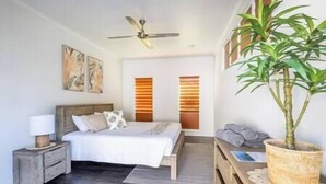 A bedroom with a bed, bedside table, lamp, ceiling fan, artwork, windows, and a potted plant.