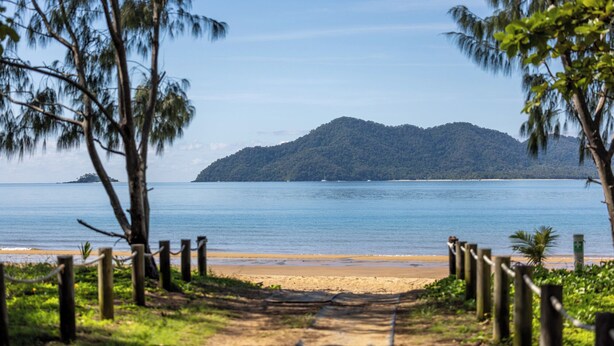 A beach path leading to a sandy beach with a wooden fence, surrounded by greenery and a clear view of a calm sea and a distant island.
