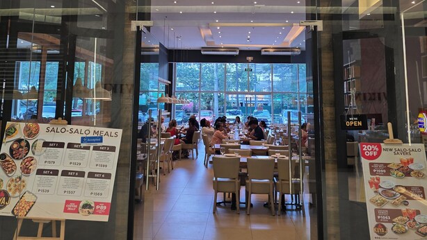 Der Eingang des Restaurants Kuya J mit einer modernen Glasfassade, einer Außenbestuhlung und freiem Blick auf die Straße.