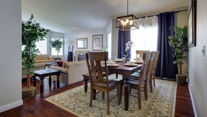 A dining area with a wooden table, chairs, and a chandelier. There is a sofa and a coffee table in the background. - Spacious Family Retreat near Botanica, Legoland, Frontwave Arena, & Beach! (Oceanside)