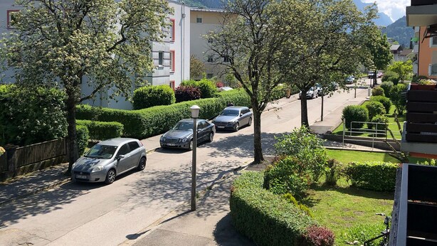 Eine Wohnstraße mit parkenden Autos, Bäumen und einem Gebäude mit rotem Fensterrahmen.