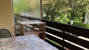 Ein Balkon mit Tisch und Stühlen, einer kleinen Ablagefläche und Blick auf Grün und Bäume.