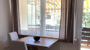 Ein Zimmer mit einem Holztisch und einem einzelnen weißen Stuhl. Ein Fenster bietet Blick auf grüne Außenbereiche und ein gelbes Gebäude.