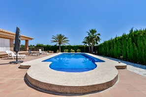 Una piscina rectangular con borde curvo, rodeada por una terraza de piedra. Hay sillas de descanso y una zona cubierta con una sombrilla.