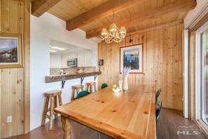 A wooden dining area with a long table, chairs, a chandelier, and a view of a mountain through a window.