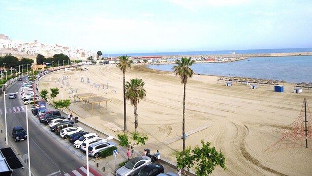 Ein Strand mit geparkten Autos und Palmen, eine Straße mit einer Fußgängerampel und im Hintergrund Meer und Gebäude.