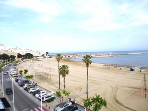 A beach with parked cars and palm trees, a road with a pedestrian crossing, and a view of the sea and buildings in the distance. - Miami Playa Sur (Peñiscola)