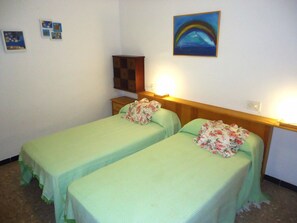 A room with two single beds, a wooden headboard, a nightstand, and a wall-mounted shelf.
