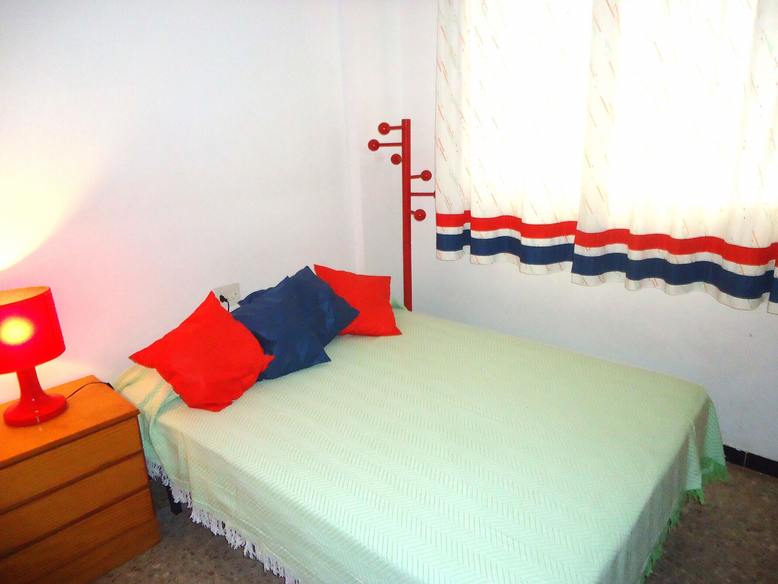 Una cama individual con un edredón verde y dos almohadas, una roja y una azul, en una habitación con paredes blancas y una ventana con cortina a rayas.
